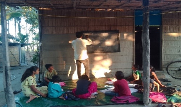 Moinuddin writing on a blackboard hung at the wall of a hut and children sitting in the front of the board looking at the content Moinuddin is writing.