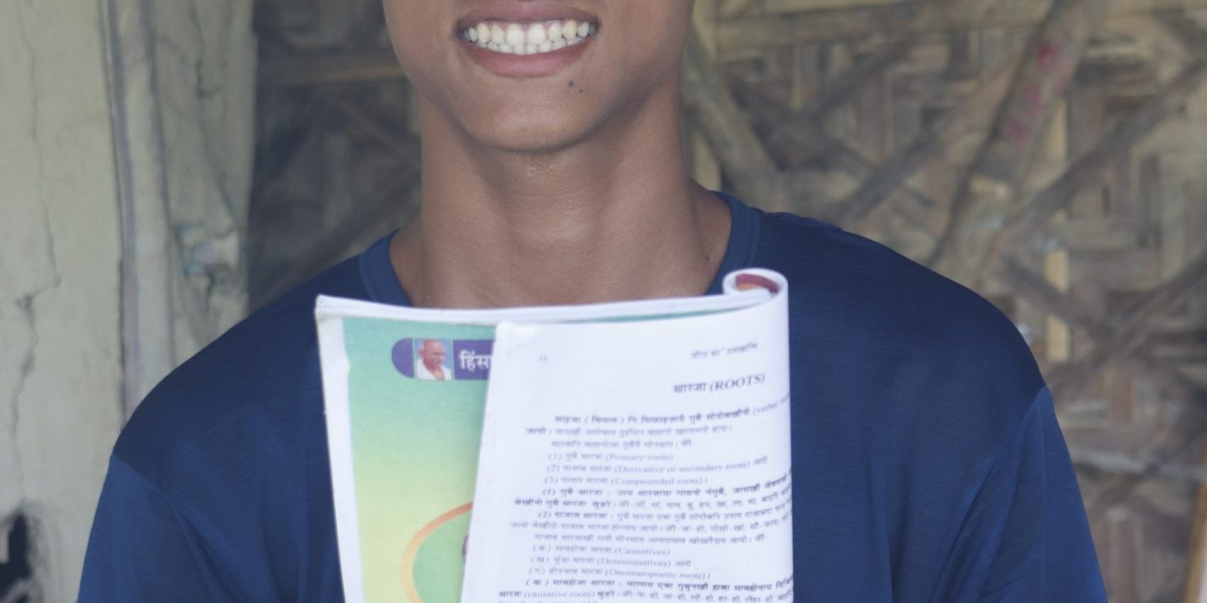 _MG_3279 Ransai in blue t-shirt holding a opened and folded school book and pen in front of him sitting in front of the door of his house and smiling at the camera.