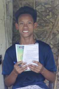 Ransai in blue t-shirt holding a opened and folded school book and pen in front of him sitting in front of the door of his house and smiling at the camera.